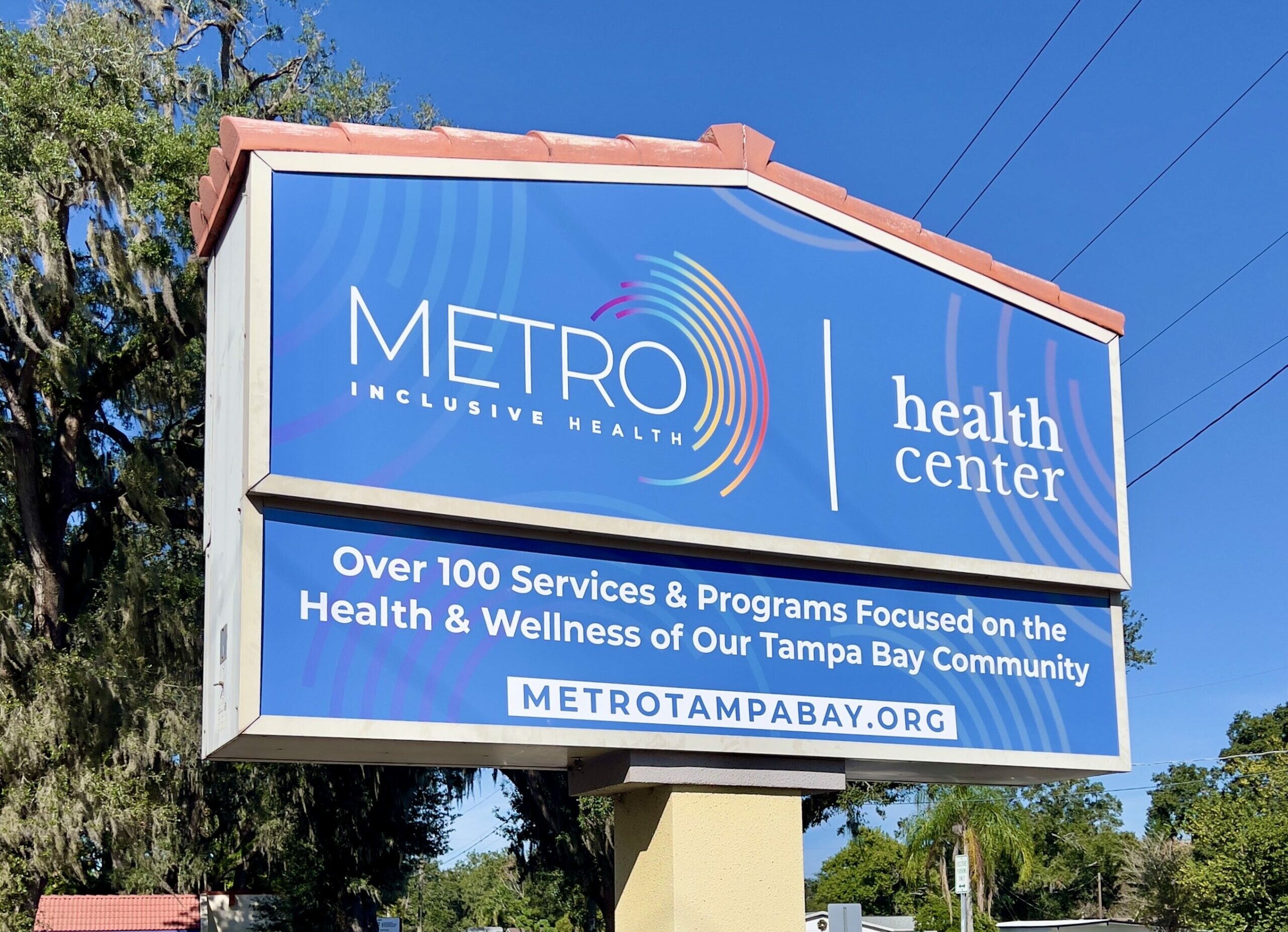 A roadside billboard for METRO Inclusive Health with a clear blue sky in the background. The sign features the METRO logo and reads 'health center' along with the message 'Over 100 Services & Programs Focused on the Health & Wellness of Our Tampa Bay Community' and the website 'METROTAMPABAY.ORG'.