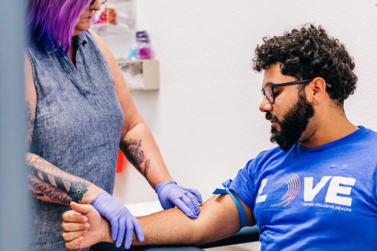 A healthcare professional with purple hair and tattoos is drawing blood from the arm of a patient wearing a blue 'LOVE Metro Inclusive Health' t-shirt. Both individuals are focused on the procedure, wearing gloves for safety, within a clinical setting.