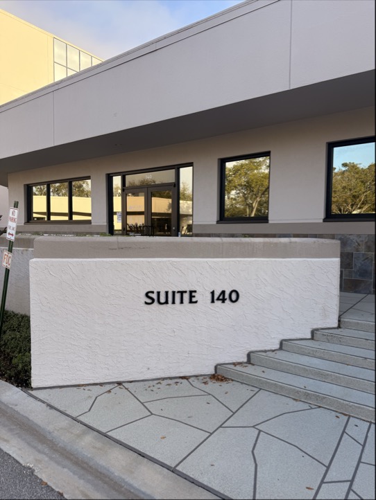 Exterior of a modern clinic building featuring a short white wall with the text "SUITE 140" next to shallow concrete steps.