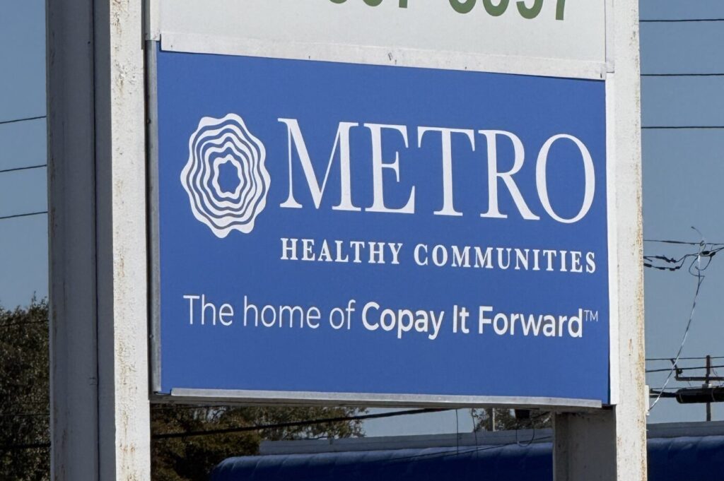 A blue outdoor sign for METRO Healthy Communities with text that reads "The home of Copay It Forward".