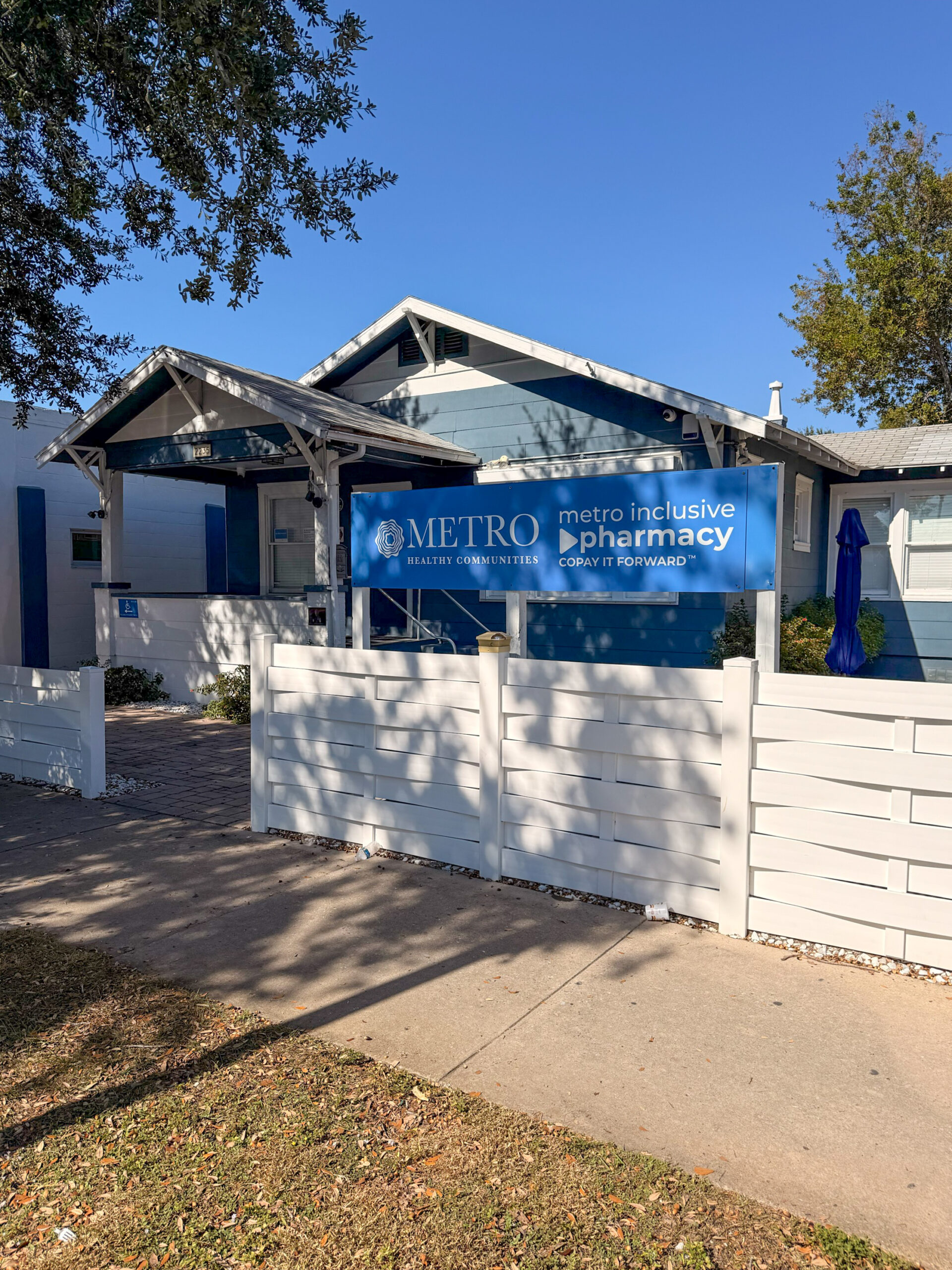 A blue and white building behind a low white fence. A large blue sign in front reads "METRO HEALTHY COMMUNITIES metro inclusive pharmacy COPAY IT FORWARD".