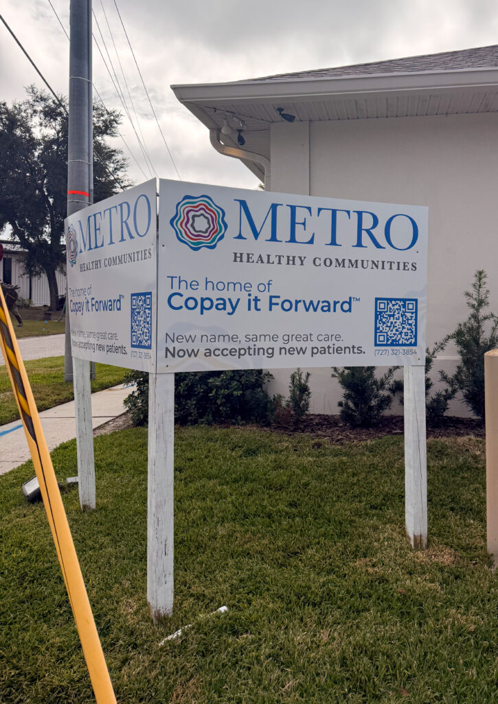 An outdoor sign on a grassy lawn for Metro Healthy Communities. The text on the sign reads, "The home of Copay it Forward. New name, same great care. Now accepting new patients," alongside a QR code.