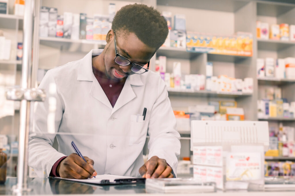 A pharmacist in a white lab coat, wearing glasses, writes notes while working in a pharmacy. Shelves stocked with various medications and products are visible in the background.
