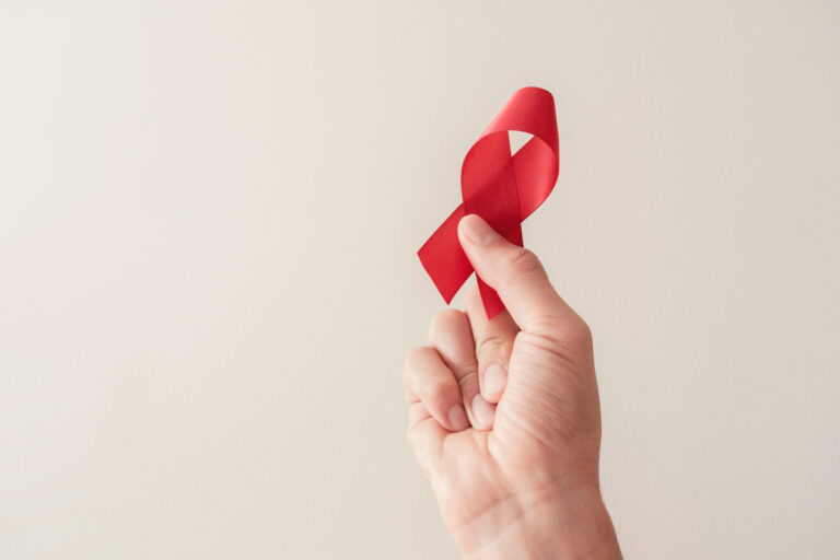 A close-up of a hand holding a small, looped red ribbon against a plain, light beige background.