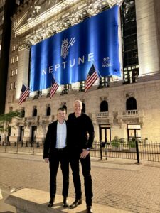 Two smiling men standing together outdoors at night in front of an illuminated historic building displaying a large blue "NEPTUNE" banner and American flags.