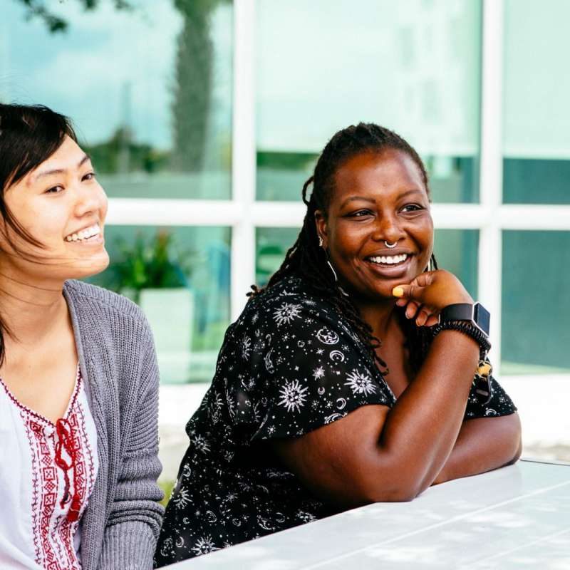 A diverse group of four people sitting around a white outdoor table, smiling and engaged in conversation.