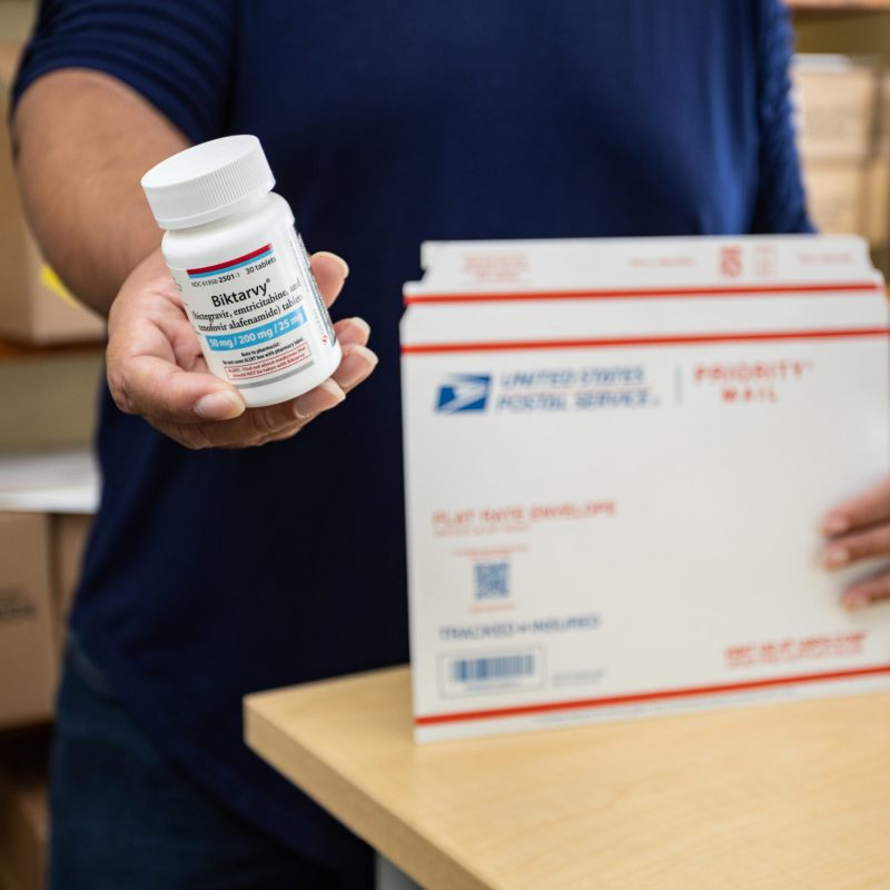 A man holding a bottle of prescription Biktarvy and a USPS Priority Mail shipping envelope.