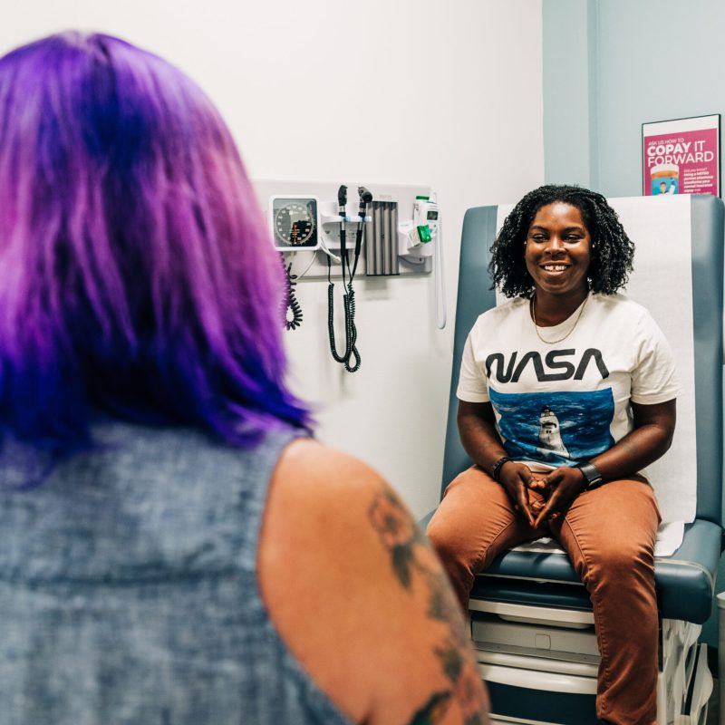 A smiling patient sitting on an exam table consulting with a healthcare provider.